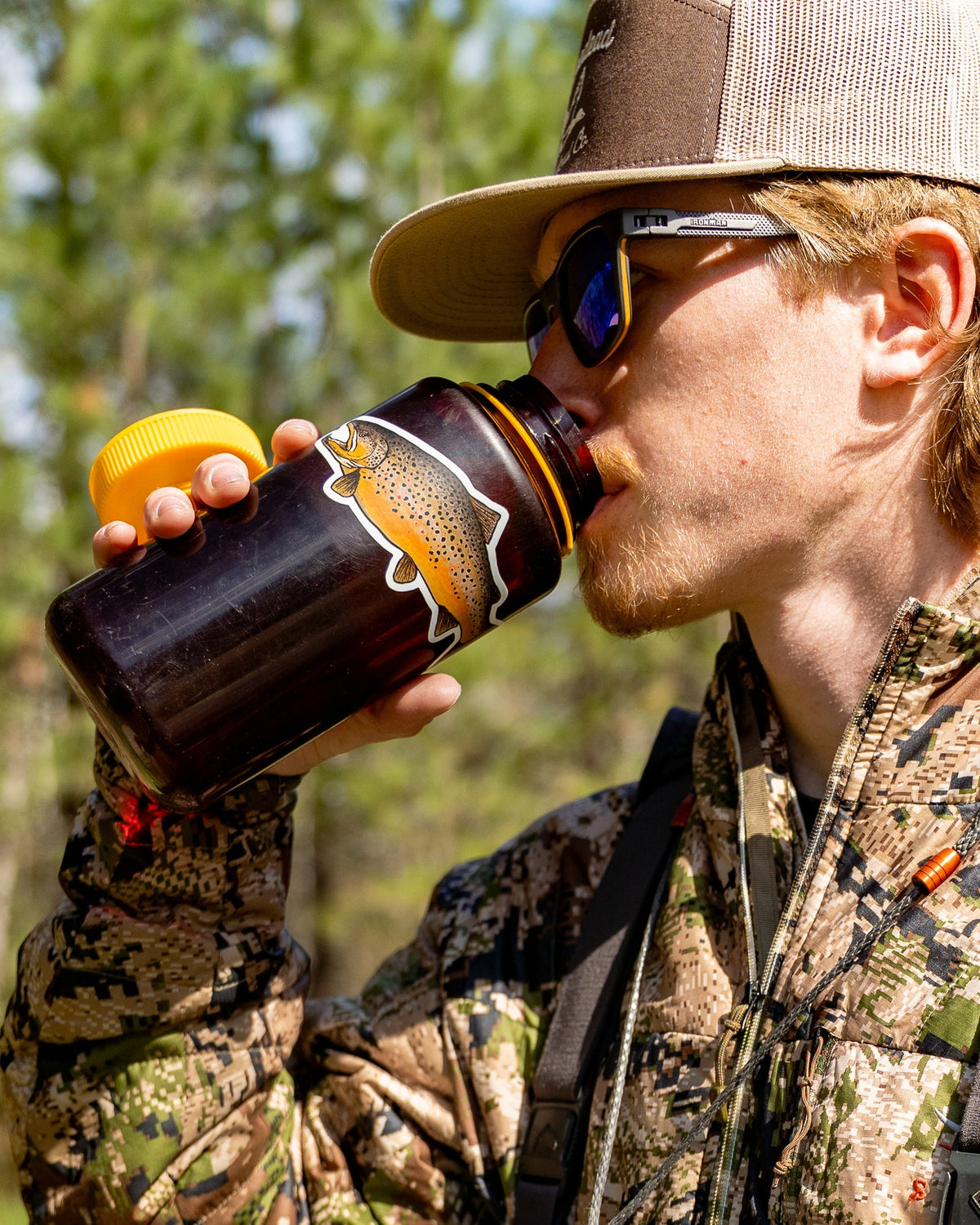 A man in camouflage hunting clothing and a cap drinks from a dark brown bottle with a yellow cap and a deer-head label, set against a blurred forest background.