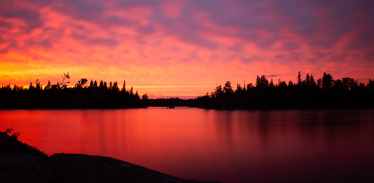 A scenic landscape photograph of a lake at sunset, featuring a vibrant orange and pink sky reflected in the calm water, with dark silhouettes of trees along the shoreline.